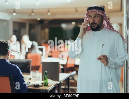 Serious Middle Eastern businessman in kandura having phone call and looking away while standing in the office Stock Photo