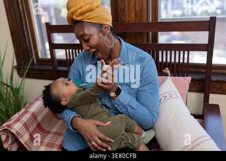 Black mother wearing yellow head wrap and long sleeve denim shirt carrying and playing with her baby boy with pacifier while sitting on wooden sofa... Stock Photo