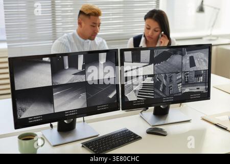 Image of two computer screens with multiple surveillance camera feeds with Black woman and mixed-race in background at monitoring and security office Stock Photo