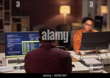 Black male programmer examining computer codes while sitting at table near mixed-race female colleague working late at night in office Stock Photo