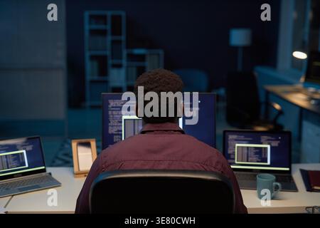 Black male programmer typing computer codes while sitting at table with laptops working late at night in office Stock Photo
