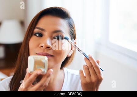 Asian woman applying eyeshadow using makeup brush while standing in living room at home Stock Photo