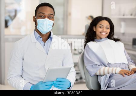 Black male dentist wearing face mask and gloves holding tablet sitting next to female patient with disposable bib ready for dental treatment in clinic Stock Photo