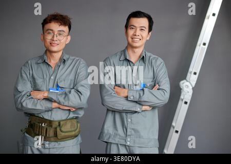 Asian male builders in uniform standing with crossed arms near metal ladder against gray wall on construction site Stock Photo
