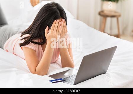Asian woman covering face with hands having financial problems while lying down on bed with laptop and credit card at home Stock Photo