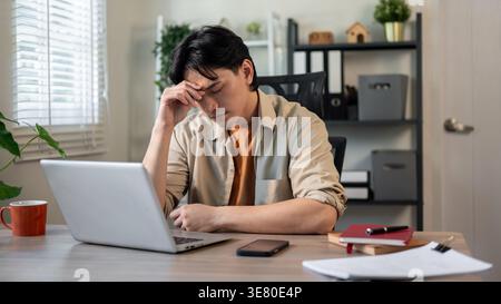 Thai businessman with closed eyes touching head suffering from headache sitting at table with laptop, cup of coffee and books in office Stock Photo