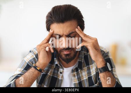 Stressed Arab man touching temples while sitting with closed eyes at table in living room at home Stock Photo