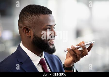 Smiling Black businessman in suit recording voice message using smartphone standing in office corridor Stock Photo
