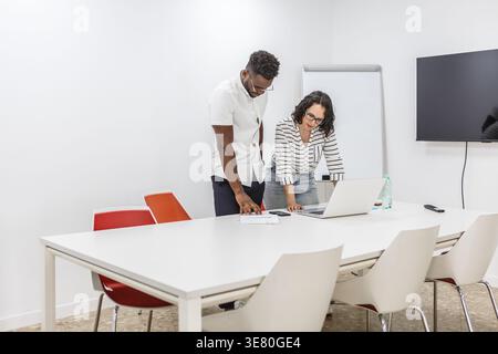 Black man and caucasian woman wearing eyeglasses standing at table and leaning over an open laptop discussing project in conference room Stock Photo
