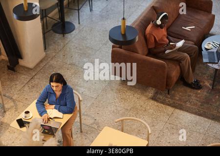 High-angle view of Mixed-race woman working on her laptop while Black man with headphones sketching on pad sitting on couch in cafe Stock Photo