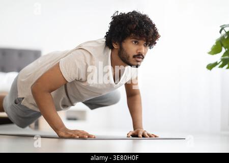 Asian man with curly hair looking away while planking on yoga mat in bedroom at home Stock Photo