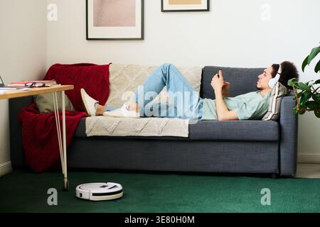Mixed-race man using smartphone listening to music with headphones lying on sofa near table in living room at home Stock Photo