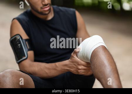 Black man wearing sportswear with armband smartphone wrapping injured knee while sitting outdoors during daytime Stock Photo