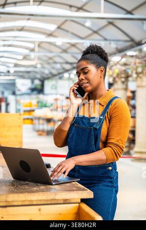 Hispanic female employee talking on mobile phone and managing orders using laptop while standing at wooden table in garden center Stock Photo