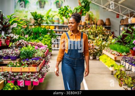 Hispanic female gardener in denim overalls smiling at plants while walking in greenhouse Stock Photo