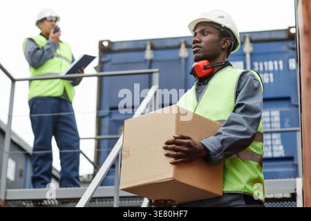 Black male worker wearing vest and white hard hat carrying box looking away walking downstairs in shipping port Stock Photo