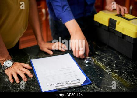 Cropped image of Asian repairman hands and female house owner leaning on table with document to fill in kitchen at home Stock Photo