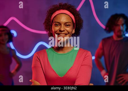 Black female teenager with headband on afro standing with folded arms in neon-lighted dark studio Stock Photo