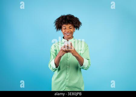 Smiling Black male with curly hair and dental braces using smartphone standing in front of blue background in studio Stock Photo