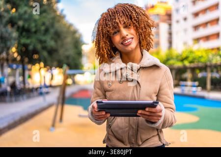 Smiling mixed-race woman with blonde twists wearing brown jacket holding tablet while standing in city park during daytime Stock Photo