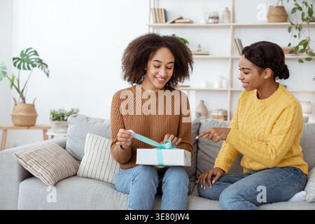 Black woman giving gift box to female friend while sitting on sofa in living room at home Stock Photo