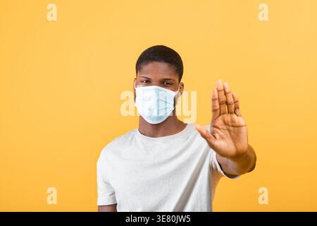 Black man with face mask gesturing stop standing against yellow background in studio Stock Photo