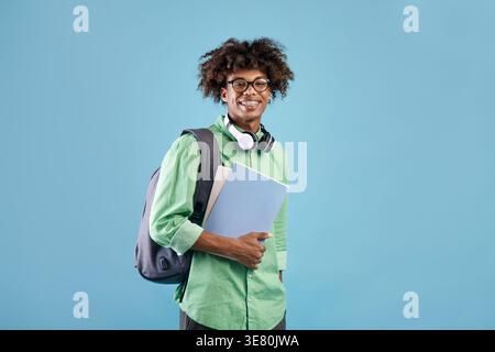 Smiling Black male student with backpack wearing eyeglasses holding books while standing sideways in front of blue background in studio Stock Photo