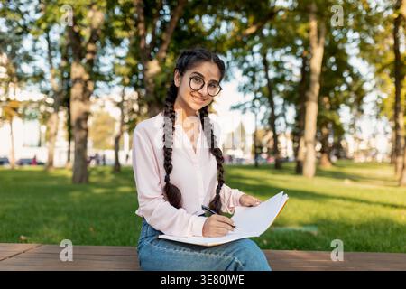Indian female student wearing eyeglasses writing on notepad sitting with crossed legs on bench in campus park during daytime Stock Photo