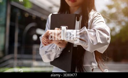 Thai business woman holding clipboard near chest looking at watch late for work standing on street in city during daytime Stock Photo