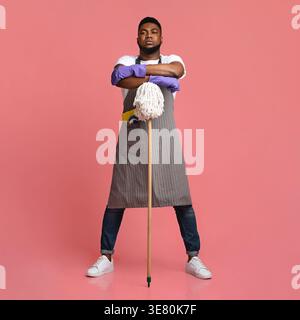 Black man with beard wearing apron and rubber gloves standing with arms crossed leaning on mop against pink background in studio Stock Photo