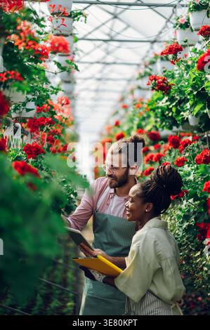 Black woman holding clipboard checking flowers with caucasian male coworker holding tablet working together in greenhouse during daytime Stock Photo