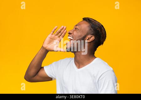 Black man in white shirt looking away shouting with hands near face standing against yellow background in studio Stock Photo