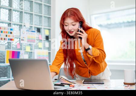 Asian woman with red hair having phone call working on her laptop while standing at table in office Stock Photo