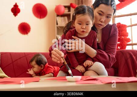 Asian mother and daughter writing with ink and brush doing traditional calligraphy while sitting on sofa with baby girl in living room at home Stock Photo