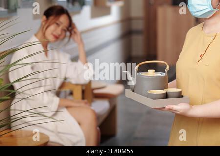 Asian female staff with face mask holding tray with pot of herbal tea and cups walking in spa Stock Photo