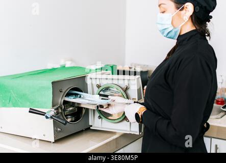 Nicaraguan female dentist wearing gloves and face mask standing at table and sterilizing tools on dental autoclave in clinic Stock Photo