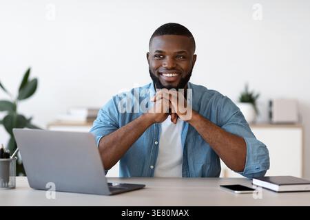 Smiling bearded Black male freelancer wearing blue long-sleeved shirt looking away leaning on clasped hands while sitting at table with laptop and ... Stock Photo