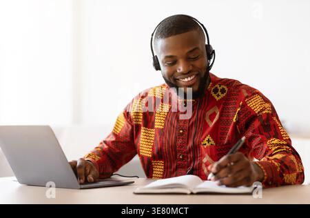 Bearded Black male freelancer wearing long-sleeved dashiki shirt writing on notebook while having videocall on laptop sitting at table in home office Stock Photo