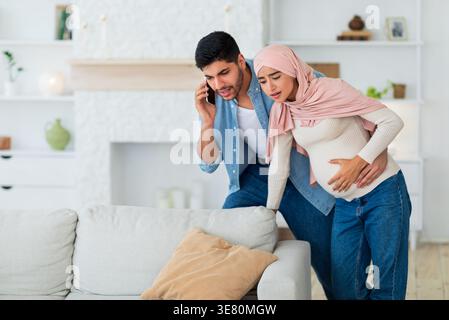 Worried Middle Eastern husband having phone call and supporting pregnant wife suffering prenatal contractions while walking in living room at home Stock Photo