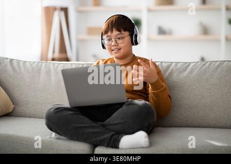 Smiling Asian boy with eyeglasses listening to music on headphones and using laptop while sitting on sofa in living room at home Stock Photo