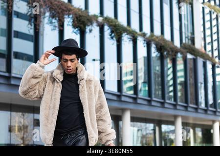 Hispanic man wearing fedora hat and fur coat looking down walking on city street during daytime Stock Photo