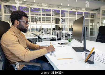 Black male architect with beard wearing brown turtleneck and eyeglasses working on blueprints for new project while sitting at desk with computer i... Stock Photo