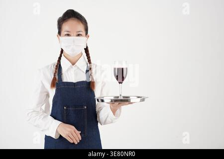 Asian waitress wearing apron and face mask holding tray with glass of red wine while standing against white background Stock Photo