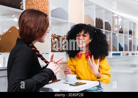 Mixed-race woman with curly hair wearing headphones around her neck having fun sitting at table and talking with caucasian female friend in cafe Stock Photo