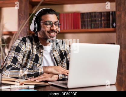 Indian male student in headphones wearing eyeglasses learning online using laptop sitting at table in library Stock Photo