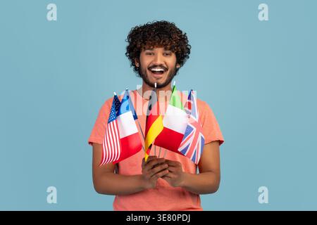 Smiling bearded Indian man with curly hair holding different national flags while standing in front of blue background in studio Stock Photo