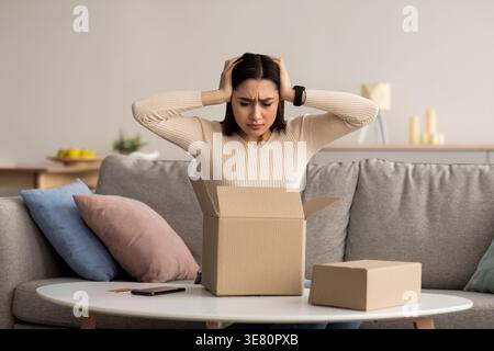 Disappointed Arab woman with hands on head looking at cardboard box having problem with her package sitting on sofa near table in living room at home Stock Photo