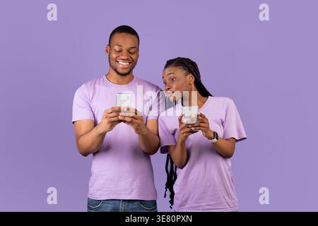 Black couple using smartphones while standing against purple background in studio Stock Photo