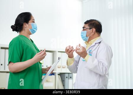 Indian male doctor wearing medical mask gesturing hands talking with Asian female nurse standing in hospital Stock Photo