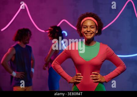 Black female teenager with afro standing with hands on waist in neon-lighted dark studio Stock Photo
